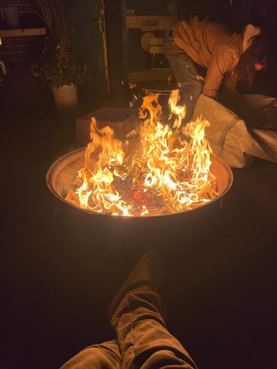 A bright fire burns in a circular pit. To the left, a person in a brown jacket and jeans sits near a green metal structure with foliage in front. Another person's legs, clad in brown pants and boots, are visible to the right, contrasting with the darkness of the background. The image is rotated, making the scene appear at an angle.