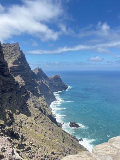 A high-angle shot shows the rocky coastline of La Aldea de San Nicolás, Spain. Sheer cliffs descend sharply to the ocean, where waves crash against the rocks and coastline. The ocean extends to the horizon with a bright blue sky. A few wispy clouds are visible. The cliffs are rugged and appear to be vegetated in places with green ground cover. The edges of the cliffs facing the water are sharp and steep. The water is a deep turquoise color closer to the coast, fading into a lighter blue further out.