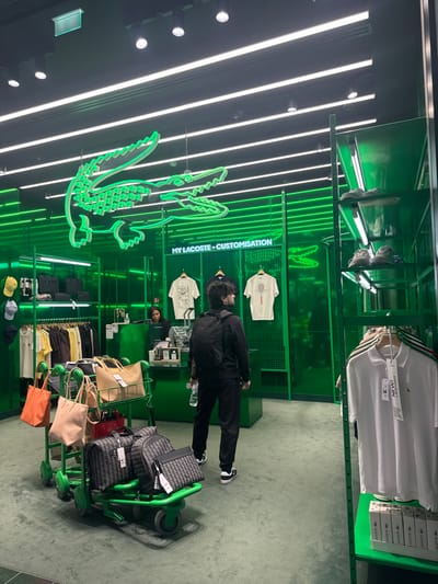 The image depicts the interior of a Lacoste retail store, likely located within a shopping area in Roissy-en-France, France, such as Charles de Gaulle Airport. The store is characterized by its vivid green color scheme, with green walls, display fixtures, and flooring. The ceiling is black, featuring multiple linear fluorescent light fixtures and recessed spotlights, providing bright illumination.
Prominently displayed on the black ceiling is a large, illuminated neon green Lacoste crocodile logo. On the back wall, above a grid display, the text "MY LACOSTE - CUSTOMISATION" is visible in white, accompanied by a smaller neon green crocodile logo.
In the middle ground, a male individual, appearing to be in his 20s or 30s with dark, curly hair, stands facing away from the camera, looking towards the back of the store. He is wearing a black long-sleeved top, black trousers, and black and white sneakers. He carries a black backpack and holds a clear plastic water bottle in his left hand.
Behind a green counter in the background, a female individual, possibly a store employee, is partially visible. She has dark hair and is wearing a dark top, attending to items on the counter.
To the left, merchandise including various colored polo shirts and t-shirts hang on racks, while shelves above display accessories like caps and black bags. The customization area features two white t-shirts with graphic designs (one abstract, one with a tennis racket motif) on hangers.
To the right, a tall, multi-tiered green shelving unit displays sneakers on upper shelves and white polo shirts with tags on lower racks. Below the shirts, stacks of white Lacoste-branded boxes are visible.
In the lower-left foreground, a green-wheeled shopping cart or luggage trolley carries several items, including an orange canvas tote bag, a beige tote bag, a smaller pinkish-beige tote bag, a red clutch, and four black/dark grey patterned luggage bags or large handbags, all with tags, indicating recent purchases.