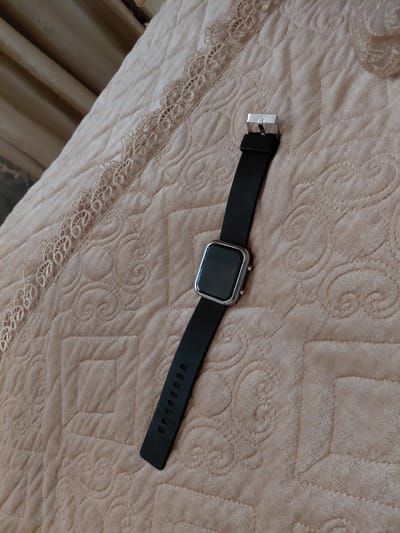A square-faced black smartwatch with a silver buckle lies diagonally on a quilted, pale pink fabric surface. The fabric has an embossed pattern of swirls and squares. A lace trim runs along the left edge of the fabric. The smartwatch is positioned with the buckle towards the upper left and the watch face towards the lower right. The watchband has a series of holes for adjustment. The scene is lit by natural light, casting soft shadows. The overall impression is one of domestic tranquility. The image is not taken in Preobrazhenskoye, Russia, as there are no specific identifiable landmarks or contextual clues to suggest this location.