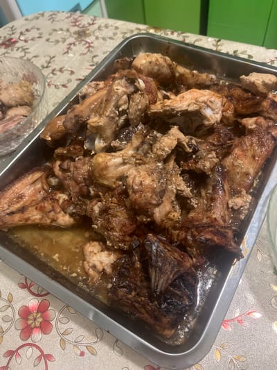 A close-up, high-angle shot shows a metal rectangular baking pan filled with cooked chicken pieces on a patterned tablecloth. The chicken is golden brown and appears to be glazed or sauced, with some pieces showing charring. The pan is brimming with chicken, making it difficult to discern individual pieces clearly. The tablecloth has a floral design with pink and red flowers and green leaves on a light beige background. In the upper left corner, a glass bowl containing more cooked chicken is partially visible. The background is slightly out of focus, showing a greenish surface and a hint of another object. The scene appears to be set indoors, likely in a dining area. There is no indication of the city of Tolsty, Russia, or any specific environmental details related to its location.