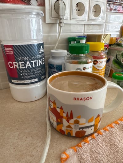 The scene depicts a countertop arrangement in Ciorescu, Moldova. In the foreground, a white ceramic mug with an orange rim holds a light brown liquid; the mug is decorated with stylized multi-colored buildings and the word "BRAŞOV" in red capital letters. To the left of the mug stands a large, clear plastic tub of "MONOHYDRATE CREATINE" dietary supplement, labeled "750 g" and "5000 MG PER SERVING." Behind these items, against a tiled white backsplash, are several smaller supplement bottles: a white bottle with "В6" visible, a clear bottle with a green cap containing gummies (labeled "FAMILY"), and another clear bottle with a yellow cap containing reddish capsules (labeled "FAMILY OMEGA 3"). A triple European-style electrical outlet is mounted on the tiles, with a white power cord plugged into the leftmost socket, trailing down onto the speckled light beige countertop. A small, light-colored textured cloth with orange trim is visible in the bottom right corner.