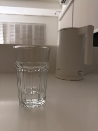 A clear glass filled with water sits on a white countertop in a room that appears to be a kitchen or dining area. Behind the glass, a white electric kettle is visible. The background shows a white wall with what appears to be closed white blinds or shutters on a window. The lighting is soft and ambient, suggesting an indoor environment. The image does not contain any information about the city of Bang Kaeo Subdistrict, Thailand, or any specific actions or interactions.