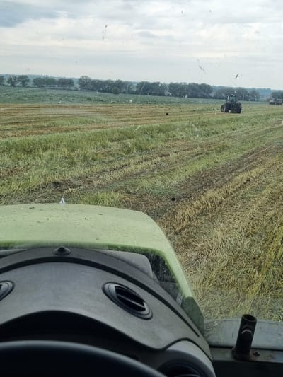 The image shows a view from inside a tractor, likely in a rural agricultural setting. The tractor is traveling through a harvested field, leaving rows of stubble. Another tractor is visible further down the field. The field is bordered by trees on the left side, and the sky is overcast. The interior of the tractor is visible, including the dashboard and steering wheel. It's unknown if this scene is in Lomnitsa, Bulgaria, based on visual evidence.