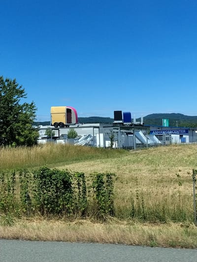 In Wörth an der Donau, Germany, a yellow and pink trailer, resembling a horse trailer, is prominently displayed on the roof of a building. To the right of the trailer, two dark blue and black rectangular structures are also situated on the building's roof. The building itself is a low-rise commercial structure with a white facade and a blue trim, featuring a sign that reads "ANHÄNGERCENTER". Adjacent to the building, a green sign with white lettering that says "StayWa" is visible. The scene is set against a backdrop of a clear blue sky and rolling green hills in the distance. In the foreground, a field of dry, golden grass stretches out, bordered by a dense green hedge. The image appears to be taken from a roadside perspective.