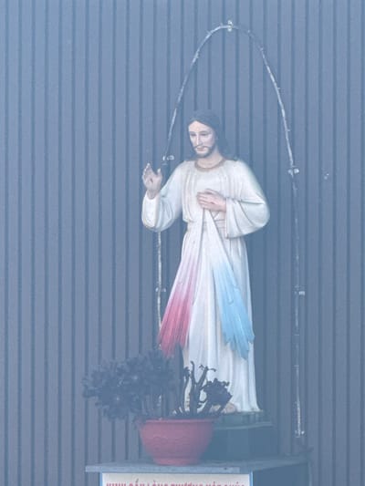 A statue of Jesus Christ is displayed outdoors in front of a corrugated metal wall. The statue depicts Jesus with his right hand raised and his left hand holding a white cloth that transitions into red and blue hues. He wears a white robe. A thorny archway made of metal frames the top of the statue. In front of the statue, to the left, is a red pot containing a dark-colored plant. The base of the statue rests on a shelf, below which a sign with text is partially visible. The overall image has a slightly hazy or misty appearance. The location is identified as Tuy Son, Vietnam.