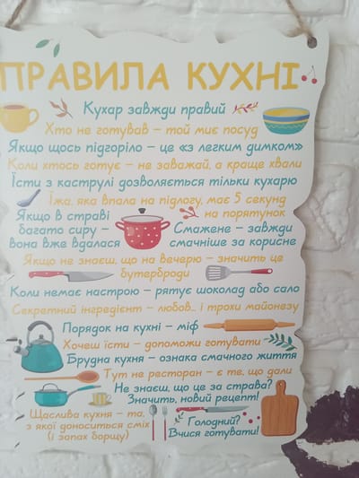 The image shows a decorative wooden sign with a list of rules for a kitchen, written in Ukrainian. The sign has a white background with colorful illustrations of kitchen items such as a teapot, a pot, a spatula, a rolling pin, a cutting board, a knife, a spoon, and a fork. There are also drawings of leaves and cherries. The text is in yellow and blue. The sign appears to be hanging indoors, possibly on a wall, as indicated by a piece of twine at the top and the blurred background which resembles a white brick wall. There is no indication of the city of Terebovlia, Ukraine, or any specific location within it. The scene is static and there are no people present.