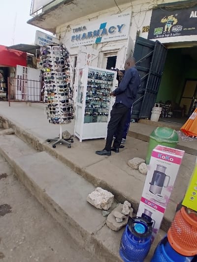 The image depicts an outdoor commercial street scene, likely in Zambia, indicated by the phone number format on a visible sign. An adult male, bald and wearing a dark blue long-sleeved shirt, dark trousers, and dark shoes, stands facing a white, glass-fronted display case filled with wristwatches. To his immediate left, a tall, rotating metal rack holds numerous pairs of sunglasses.
Behind these displays, a building features two prominent signs: "TINNA PHARMACY" with the telephone number "09672448283," and a partially obscured sign for "TINNA PHARMACEUTICAL CHEMICALS." To the right of the pharmacy entrance, a dark banner with a muscular graphic advertises "FMN BAGGO" and reads "...body bags it better." The ground consists of concrete steps and unpaved areas. On the lower ground level to the right, several small blue and orange gas cylinders, some with attached burners, are present. A pink and white box labeled "KoKo VACUUM FLASK" stands upright on the upper concrete platform. Two green cylindrical trash bins are also visible near the gas cylinders. Scattered rough rocks lie on the concrete surfaces. Another person's leg, clad in blue trousers, is partially visible behind the main figure. No visual information within the image identifies or locates Kungulu School.