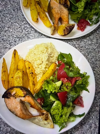 This overhead shot captures two plates of a prepared meal, likely served in an indoor setting with a grey, speckled countertop as the surface. The foreground plate is the primary focus, showcasing a balanced meal.

On the main plate, a piece of cooked fish, possibly salmon or mackerel, is prominently displayed, showing golden-brown skin and flaky flesh. Alongside the fish are several golden-yellow potato wedges, appearing baked or roasted. A substantial mound of light-colored, possibly brown, rice or bulgur is placed next to the potatoes. Completing the meal is a fresh green salad, composed of mixed leafy greens, diced red tomatoes, and sliced green cucumbers. The salad appears lightly dressed.

Partially visible in the upper left corner is a second plate, presenting a similar meal composition with another piece of cooked fish, potato wedges, and a green salad with tomatoes. A charred lemon wedge is visible near the fish on this second plate, suggesting it's also part of the serving.

The overall scene depicts a homemade or restaurant meal, neatly arranged on white, round plates. There are no people visible, and no visible text. The image provides no specific time of day or weather cues beyond an indoor setting. While no visual cues directly confirm the location, the user specifies the context as Ros Na Rí, Ireland.