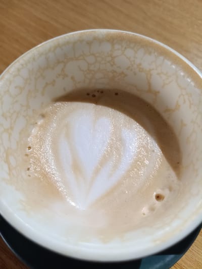 A close-up, overhead shot shows a white ceramic cup containing the remnants of a latte. The inner rim of the cup is stained with a light brown residue, forming an abstract, web-like pattern. The remaining liquid in the cup is frothy and a light brown color, with a visible heart-shaped latte art design in the center. A few small air bubbles are present on the surface of the liquid. The cup rests on a dark saucer, which is partially visible. The background is a wooden surface, likely a table. The scene is devoid of any people or animals. This image is not associated with the city of Buzău, Romania, as there are no elements in the image that indicate a specific geographical location.