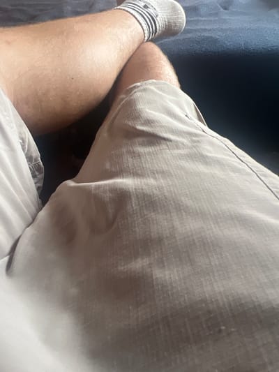 This image shows a close-up, over-the-shoulder view of a person's legs and torso. The person is wearing light-colored shorts with a textured fabric and a pair of gray athletic socks with black stripes. The legs are bare and appear to be lightly tanned, with visible hair follicles. The background is dark and out of focus, with a suggestion of a dark fabric surface, possibly a couch or chair. The lighting appears to be natural, suggesting daytime. There is no visible text in the image. The framing focuses on the lower body, with the upper part of the shorts dominating the right side of the frame. The scene is casual and relaxed, with no specific activity or event clearly depicted. The overall impression is one of a person resting or sitting in a comfortable environment.