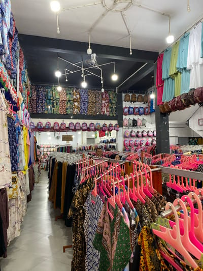 This media file depicts the interior of a retail store, likely a fabric shop or a boutique selling clothing and accessories, located in Lamteumen Barat, Indonesia. The scene is brightly lit, suggesting it is daytime, and the store appears well-stocked and organized.
The primary subjects are rows of various fabrics, garments, and accessories displayed on shelves, racks, and tables. Prominently featured are stacks of colorful fabrics with intricate patterns, as well as many pink plastic hangers holding clothing items. Several rows of backpacks, in various colors and designs, are neatly arranged on shelves, indicating a section for children's items or fashion accessories. Some handbags or purses are also visible hanging from racks.
The overall activity is the display and sale of merchandise. There are no people directly visible engaging in the shopping activity, but a few reflections in mirrors or glass surfaces might hint at the presence of staff or customers not clearly in frame. The setting is a commercial space with lighting fixtures and shelving units.
Notable details include the sheer volume and variety of textiles, suggesting a wide selection for customers. The use of colorful displays and organized shelving creates a vibrant and inviting atmosphere for shoppers. The presence of Indonesian text on a sign in the background, though illegible in this resolution, confirms the location context. The image captures a typical retail environment focused on apparel and fabrics.