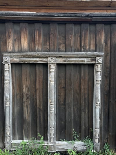 An aged, weathered wooden frame, likely for a double window, leans against a vertical wooden wall in the city of Nogornyi, Russia. The frame, painted a faded grey, shows signs of peeling paint and wear. It is ornately carved with decorative elements, including circular motifs at the top corners and stacked cylindrical shapes along the vertical posts. The wooden wall behind it is constructed of dark, rough-hewn planks, showing a distinct grain pattern and varied tones of brown and grey, suggesting it has also been exposed to the elements for a considerable time. Greenery, including grass and small vines, grows at the base of the wall and around the lower portion of the frame. The overall scene conveys a sense of age and abandonment.