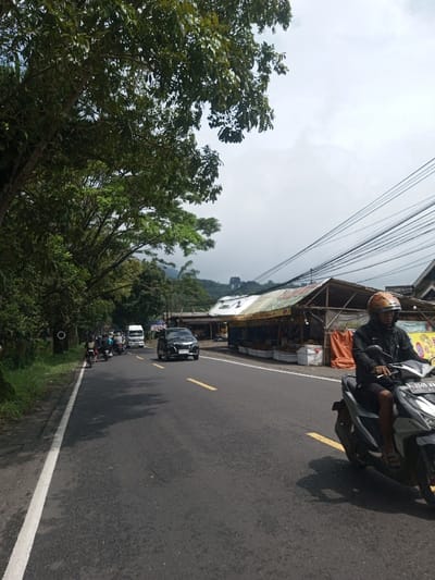 The image depicts a roadside scene in the city of Mahawilachchiya, Sri Lanka, under an overcast sky. A two-lane asphalt road, marked with a solid white line on the left edge and dashed yellow lines in the center, extends from the foreground into the midground.
In the immediate foreground on the right, a person is operating a silver-grey scooter (motorcycle). The rider wears a dark long-sleeved top, dark shorts, sandals, and a light brown full-face helmet. The scooter's rear license plate, partially visible, reads "P 3345 VKG".
On the left side of the road, dense green foliage and trees line the shoulder. Further down the road, a line of traffic is visible, predominantly consisting of motorcycles, some carrying riders. A white utility truck follows behind the motorcycles. Traveling in the opposite direction, a black passenger car is positioned closer to the center of the road.
On the right side of the road, opposite the dense greenery, a series of makeshift market stalls are present. These structures feature wooden frames, a mixture of corrugated metal and tarpaulin roofs (green, yellow, and orange), and displays of various goods, possibly produce, arranged in white crates. Multiple black electrical wires are strung overhead, crisscrossing the sky above these stalls.
In the background, distant hills or mountains, partially covered in vegetation, are visible beneath the cloudy sky. Some lighter rock formations or cleared areas are identifiable on the distant slopes.