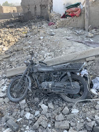 The image depicts a scene of extensive destruction in Spin Boldak, Afghanistan, characterized by widespread rubble and damaged structures under direct sunlight. In the foreground, a dark, heavily soiled, and partially burnt Honda motorcycle lies on its left side amidst a dense field of light gray and beige debris, comprising broken bricks, concrete fragments, and soil. The motorcycle's seat is torn, and wires are visible near its engine block.
The mid-ground features a large expanse of similar rubble, with a long, rectangular concrete beam resting horizontally across the debris. Further back, a patterned red and brown rug or blanket and a green cushion or blanket are visible on a mound of debris. A shovel or pickaxe is partially visible among the rubble.
The background reveals the remnants of severely damaged buildings. Partially standing walls, constructed from mud-brick and possibly concrete, show exposed interiors and collapsed sections. One structure, forming a corner, has a stack of what appear to be cushions or blankets piled atop a low wall. No individuals are visible in the scene, and there are no discernible actions or interactions occurring. The environment conveys desolation and the aftermath of significant structural damage.