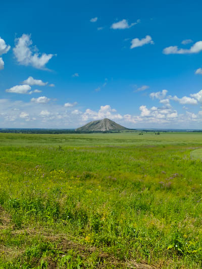 A vast, green, grassy field dominates the foreground and middle ground of this image, stretching towards a distant, conical hill. The hill, likely a natural formation or an ancient burial mound, appears light brown and grey, its slopes textured by time and weather. A dense line of trees forms a dark green horizon behind the field, separating it from a hazy, pale blue sky. The sky is dotted with fluffy white clouds, some small and wispy, others larger and more defined, casting subtle shadows across the landscape. The overall impression is one of serene, open countryside under a bright, sunny sky. The image is a landscape shot with no people or animals present.