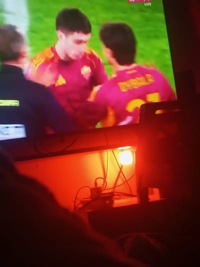 A television displays three soccer players in red uniforms on a green field. The player in the center faces forward, while the player on the right has "DYBALA" and the number "21" on his back. A person in dark clothing stands on the left. Below the television, a shelf contains electronic devices and various cables. An orange light illuminates the shelf. The scene appears to be viewed from a low angle, with part of a person's body visible in the foreground, suggesting they are seated and watching television.