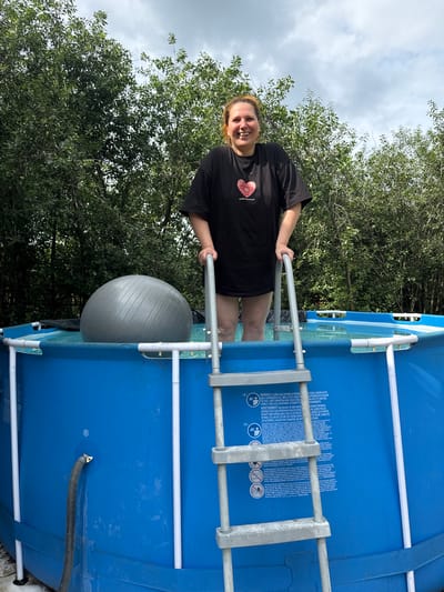 An overhead shot captures a woman with light brown hair, wearing a black t-shirt with a red heart graphic and text that reads "MADE IN RUSSIA", standing in a blue, above-ground swimming pool. She is smiling and looking at the camera. Her legs are submerged in the pool water. A grey exercise ball is partially submerged in the water next to her. A silver ladder with grey rungs extends into the pool, and the woman's right hand is resting on one of the ladder's rails. The pool is surrounded by green trees and vegetation under a cloudy sky. The scene appears to be in a backyard in the village of Khokhryaki, Russia.