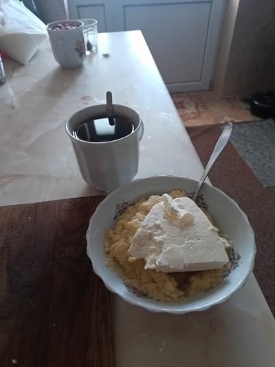 A close-up, high-angle shot shows a breakfast scene on a table in a home located in Suklieia, Moldova. In the foreground, a white bowl with floral accents contains a serving of what appears to be a creamy grain porridge topped with a substantial slab of white cheese. A silver spoon rests inside the bowl, partially submerged in the porridge. To the left of the bowl, a white ceramic mug holds dark liquid, likely coffee or tea, with a spoon resting inside. A wooden cutting board is visible to the left of the bowl, and a patterned rug can be seen in the background. Other household items, including a bag of sugar and another mug with red contents, are present on the table, suggesting a domestic setting. The lighting is natural, indicating daytime.