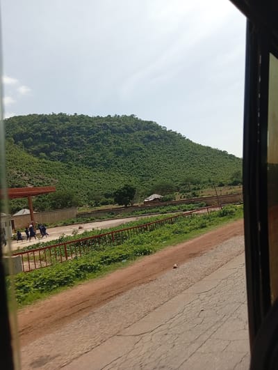 This is a photograph taken from inside a vehicle, looking out at a roadside scene. The dominant feature is a lush green, heavily vegetated hill or mountain that occupies the upper half of the image. The hill is densely covered with trees and shrubs. In the foreground, a dirt road runs diagonally from the lower left to the lower right, parallel to a paved road with visible cracks. A red metal fence runs along the edge of the paved road, separating it from a strip of green vegetation.
To the left of the paved road, there is a concrete wall with a structure attached, possibly a small building. In front of this structure, under a reddish-brown overhead canopy supported by pillars, a group of five children are walking or running. They appear to be wearing dark blue uniforms. Their interaction is unclear, but they are moving in the same direction. Beyond the children and the structure, there are more trees and a small, pitched-roof building. The sky is overcast with pale clouds. The overall scene depicts a rural or semi-rural environment with a natural landscape and signs of human activity. The location is identified as Rimin Ziem, Nigeria.
