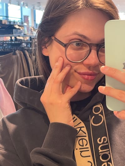 A fair-skinned woman with shoulder-length dark hair is taking a selfie indoors, likely in a retail environment. She wears round-framed glasses and a dark gray hoodie, with visible lettering that reads "Calvin Klein Jeans." The background shows clothing racks and other merchandise, suggesting a clothing store setting. The location is not discernible as Keszthely, Hungary from visual elements.