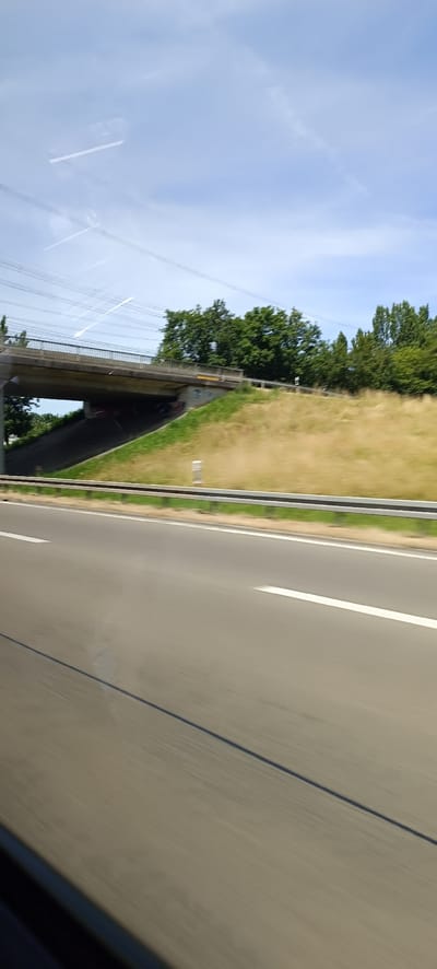 The image depicts a roadside view, likely from a moving vehicle, featuring a highway overpass and surrounding greenery under a partly cloudy sky. The overpass, constructed of concrete, spans across a grassy embankment. The embankment is covered in dry, yellowish grass on the upper portion and greener grass on the lower slope leading to a dark underpass. Some graffiti is visible on the concrete supports of the overpass. Trees with dense green foliage line the top of the embankment. Power lines are strung above and to the left of the overpass. The foreground consists of a multi-lane asphalt road, indicated by white lane markings and a dark shadow stretching across it, suggesting a sunny day. A metal guardrail separates the road from the embankment. The sky is a light blue with scattered white clouds, and a faint contrail is visible. The scene appears to be located in a suburban or rural area with some infrastructure.