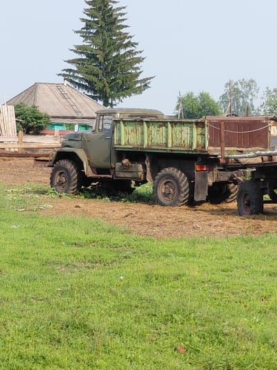 An exterior, full shot captures an olive green, vintage truck with a trailer parked on a dirt and grass area. The truck is positioned facing right, with its front slightly obscured by shadows. The truck features large, rugged tires and a weathered, green cargo bed. The trailer behind it is also a rusty brown color and appears to be carrying some unseen cargo.

In the background, a large, dark green evergreen tree stands prominently. To the left of the tree, a house with a grey, corrugated roof is visible, with some wooden structures and greenery surrounding it. The sky above is a pale, hazy blue. The overall scene suggests a rural or semi-rural setting, likely in the city of Novosyolovo, Russia, given the context. There are no people present in the image, and the environment appears to be outdoors on a clear day.