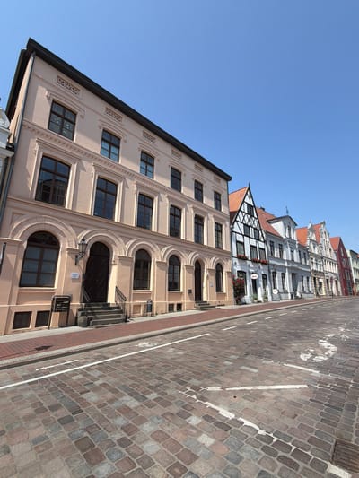 This is a low-angle, wide shot of a cobblestone street in the historic town of Wismar, Germany, on a clear, sunny day. The street curves gently to the right, lined with a row of colorful, historic buildings. The foreground is dominated by the textured cobblestones of the street, with white painted lines indicating traffic lanes and a bicycle symbol.
The buildings on the left are predominantly light-colored, with arched windows and decorative architectural details. The first building on the left is a light peach color with brown framed windows and an ornate entrance with a lamp. Further down the street, the architecture becomes more varied, with half-timbered buildings in black and white, and other structures with red tiled roofs. Some buildings have flower boxes in the windows. The sky is a bright, cloudless blue. There are no people visible in the image. The scene is static and appears to be captured from street level, looking down the length of the street.
