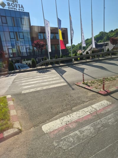 The image shows a street scene in Fălticeni, Romania, on a sunny day. In the background, the modern glass facade of the "Angel Sofia Hotel" is visible, with its name and star rating displayed prominently. Several flags are mounted on poles in front of the hotel, including the Romanian national flag. The foreground features a pedestrian crossing on an asphalt road. The crossing has white stripes, some of which are faded and marked with pink paint. Red and white striped bollards are present on a small paved area adjacent to the crossing, which is bordered by red and white curbs. Green grass and small trees are visible along the side of the road and near the hotel. A white van is parked in front of the hotel. The sky is clear and blue.