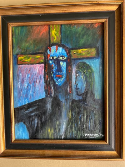 The image shows a framed oil painting of two figures in front of a cross. The figure in the foreground is larger and more detailed, with a blue face, red lips, and eyes that stare forward. The figure's body is dark and appears to be cloaked. To the right of the foreground figure is a smaller, profile view of another figure, also with blue skin and dark hair. Behind them, a large, abstract cross is depicted with bold strokes of yellow, red, and green. The background is a mix of blues and pinks. The painting is signed "L. Janssen '96" in the lower right corner. The painting is framed in a black and gold frame, which is hanging on a light brown wall. There is no information in the image to determine the location in the city of Denderleeuw, Belgium.