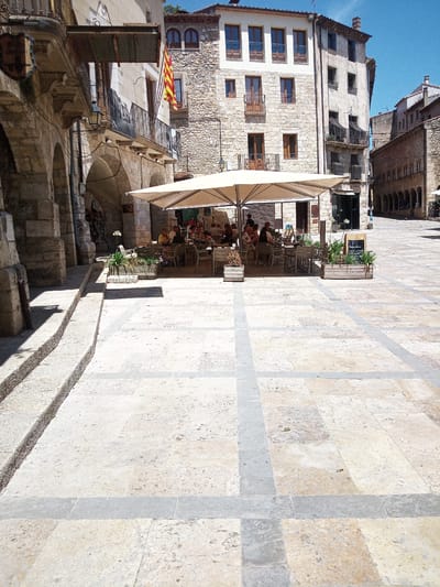 The image depicts a sunlit public square in Besalú, Spain, paved with large, light-colored stone tiles arranged in a grid pattern with contrasting grey inlays. In the mid-ground, an outdoor cafe operates under a large, cream-colored umbrella, providing shade for several tables and chairs. Patrons are seated at these tables, engaged in conversation and enjoying their meals.

To the left of the cafe, an arched stone walkway leads into a building, with a shield-like sign hanging above. A Catalan flag, with red and yellow stripes, is prominently displayed on a balcony of a stone building to the left. The buildings surrounding the square are constructed from stone, featuring traditional architectural elements such as balconies with wrought-iron railings and wooden window frames.

In the foreground, a stone staircase descends into the square. The overall scene conveys a lively atmosphere in a historic European town, with architectural elements typical of medieval Spanish towns. The sky is a clear, bright blue.
