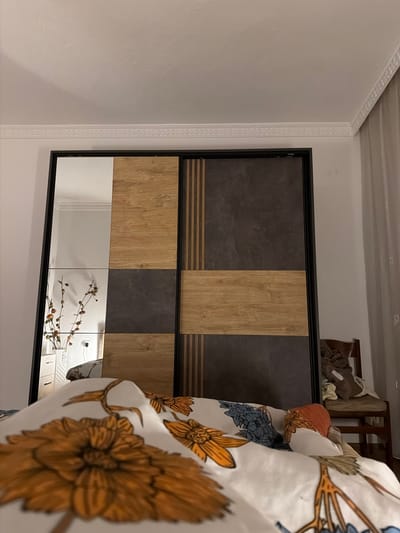 The image shows a bedroom with a large sliding door wardrobe in the center. The wardrobe has a combination of wood-grain and dark stone-effect panels, with a mirrored section on the left. A bed with floral patterned bedding is in the foreground, partially obscuring the bottom of the wardrobe. A white vase with dried branches is visible in the mirrored section of the wardrobe, reflecting a white wall with a decorative molding. To the right of the wardrobe, a wooden chair is laden with clothes. The overall impression is that of a tidy, yet lived-in bedroom. This scene is located in a bedroom in Zmeitsa, Bulgaria.