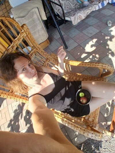 A woman lies on a wicker lounge chair outdoors. She has brown hair and is wearing a black dress. Her legs are crossed, and a small bowl of two apples, one red and one green, rests on her lap. The chair is on a paved patio with a checkered pattern of beige and brown tiles. In the background, a piece of furniture with a patterned cushion and a metal frame is visible, along with gravel and shadows cast by trees. The overall scene suggests a relaxed outdoor setting in a residential area.