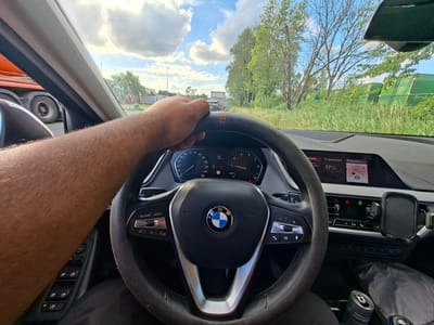The image shows a person's hand gripping the steering wheel of a BMW car. The car is on a road, with a large orange truck visible to the left and green containers in the distance to the right. The dashboard and center console of the car are also visible, including a digital display. The scene appears to be an outdoor environment, possibly a highway or a road in an industrial area. The sky is partly cloudy with blue sky visible. The location is indicated as Boom, Belgium.