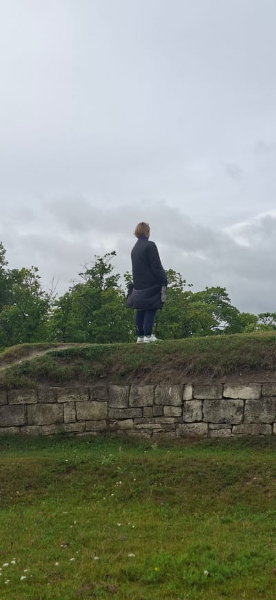 An overcast day in Kuressaare, Estonia, as a woman stands on top of a weathered stone wall. She is facing away from the camera, looking towards a lush green landscape of trees and a cloudy sky. The woman is wearing a dark quilted jacket and dark pants, with white sneakers. A dark bag is slung over her shoulder. The stone wall, constructed from rough-hewn rectangular blocks, forms a prominent horizontal line across the middle of the frame, separating the grassy foreground from the elevated area where the woman stands. The grass in the foreground is a mix of green and brown hues, dotted with small white and purple wildflowers. The trees in the background are densely packed and appear to be deciduous, their leaves a vibrant green. The sky is a uniform grey, with visible cloud formations suggesting a lack of direct sunlight.