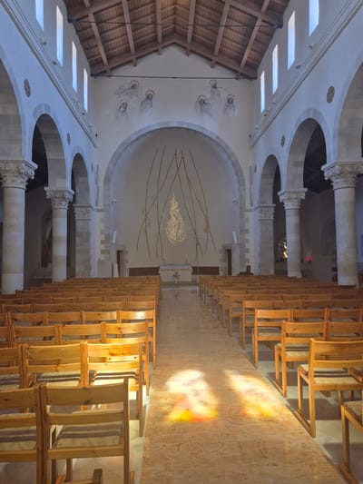 This is a wide-angle interior shot of the Church of the Annunciation in Abu Gosh, Israel. The church features a nave lined with rows of simple wooden chairs facing an altar at the far end. Sunlight streams through high arched windows, casting colorful patterns on the floor. The walls are adorned with religious artwork, including a depiction of angels and a golden mosaic. The architecture incorporates stone columns and arched doorways. The scene is empty of people.