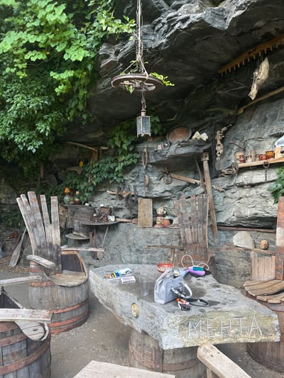 The image depicts an outdoor seating area carved into a rock face, likely located in Golyamo Belovo, Bulgaria, given the context. The setting is rustic, with a stone table supported by two wooden barrels. On the table, there are several items including two packs of cigarettes, a glass, a grey handbag, a pink electronic device, and a white electronic device. To the left of the table is a unique chair made from a wooden barrel with a tall, slatted wooden back. To the right of the table, another wooden chair, also constructed from barrel components, is visible. The rock face behind the seating area is adorned with various rustic tools and artifacts, suggesting a historical or themed display. Lush green ivy grows down from the top of the rock face, adding a natural element to the scene.