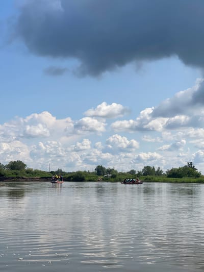 An aerial drone shot captures a wide, calm river flowing through a verdant landscape. The sky is a mosaic of bright blue and dramatic, dark grey clouds, suggesting an approaching storm. On the water, two rafts are visible. The closest raft, positioned in the middle of the frame, carries a group of people wearing life vests and holding paddles, actively engaged in kayaking. Further in the distance, a second raft, also populated by individuals in life vests, is further to the right. The banks of the river are lined with lush green trees and foliage. In the background, a few utility poles and the outline of a small aircraft can be discerned near the treeline, indicating proximity to a populated area or airfield.