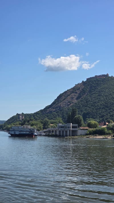 A wide, calm body of water, likely the Danube River, occupies the foreground. The water is choppy with small waves reflecting the bright sunlight. In the middle ground, a large, multi-decked riverboat is docked alongside a modern building with arched windows and a covered walkway. The boat appears to be a passenger vessel with multiple cabins and windows. Behind the riverboat and building, a lush green hillside rises steeply, covered in dense trees. At the very top of the hill, a medieval castle ruin is visible, with stone walls and towers silhouetted against the clear blue sky. A few small, white clouds are scattered across the sky. The scene captures a view of Visegrád, Hungary, showcasing its natural beauty, historical landmarks, and riverine transportation. There are no people clearly visible in the image.