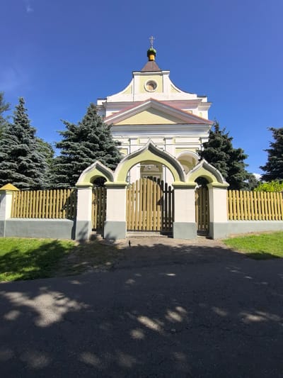 The image shows a bright, sunny day in a rural area, likely in Шчорсаўскі сельскі Савет, Belarus. The foreground is an asphalt road with shadows cast by trees. A light-colored fence with a yellow picket top runs across the mid-ground, separating a patch of grass from a building. The building, which appears to be a church or a similar religious structure, is painted yellow and white with a red roof. It features a green dome topped with a cross. An arched gateway with decorative yellow and white trim provides access through the fence. Tall evergreen trees are visible behind the fence and on either side of the building. The sky is a clear, bright blue.