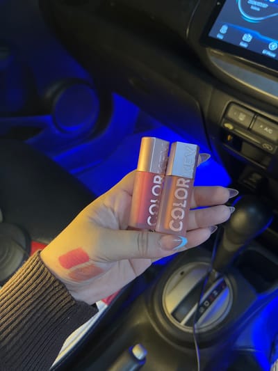 In a car interior illuminated by blue ambient lighting, a person's hand displays two lip glosses with "COLORKEY" branding. Swatches of pink and nude lipstick are visible on the back of the hand. The car's gear shift and dashboard are in the background, suggesting the vehicle is stationary. The location of Beuringen, Indonesia is not depicted in the image.