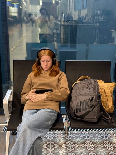 The image captures a young person resting on a black public bench, likely in a waiting area such as an airport or train station. The person, appearing to be a young female with reddish-blonde hair, is lying on her side with eyes closed, suggesting she is sleeping or deeply resting. She wears black over-ear headphones, a light brown hooded sweatshirt, and light blue jeans. A black smartphone or small tablet is held in her clasped hands, resting on her stomach.
To her upper right, on an adjacent seat, are two bags: a light brown woven straw bag and a grey backpack with a prominent, stylized "NIKE" logo visible on its side in a darker grey. The floor to the right of the seating features decorative patterned tiles in shades of grey, white, and brown/blue. A large window occupies the left side of the frame, showcasing blurry reflections of an indoor environment, including what appears to be a distorted reflection of another person or structure. A white charging module with USB ports is attached to the seating structure beneath her feet. The lighting suggests it is daytime. The scene conveys an atmosphere of quiet waiting or travel.