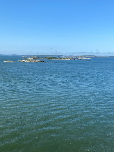 A wide shot captures a serene, sun-drenched seascape with a clear blue sky dominating the upper portion of the frame. The vast expanse of the ocean, rendered in shades of deep blue and green, fills the majority of the image, with gentle ripples creating a textured surface. In the distance, a scattered archipelago of rocky islands, sparsely dotted with vegetation and some structures, emerges from the water. A navigational buoy, painted in white and green, stands prominently near the center-left of the image, marking a channel or hazard within the waterway. The overall scene suggests a tranquil day in the archipelago of Gothenburg, Sweden, likely viewed from a boat or elevated vantage point.