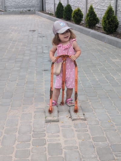 In Perliavka, Ukraine, a young girl wearing a pink polka-dot romper and a grey baseball cap stands on a cobblestone walkway. She is holding onto an orange metal barrier, which appears to be a parking bollard or similar street furniture. The barrier is weathered and shows signs of rust. The girl has a small, woven bag hanging from her shoulder. She is wearing pink clogs. Behind her, a row of small evergreen trees is visible, backed by a textured concrete wall. The overall environment suggests an outdoor, possibly residential or public space.
