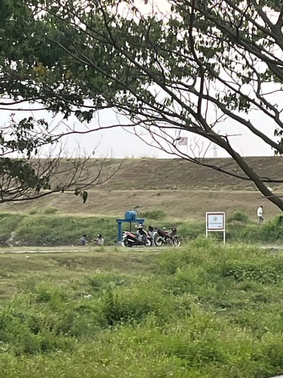 The image depicts an outdoor scene with a large tree in the foreground, its branches partially obscuring the view of the background. The middle ground features a grassy area with several people and motorcycles. A blue structure, possibly part of a water pump or filtration system, is visible near the motorcycles. Further in the background, there's a raised embankment covered in dry grass. A white sign with a red symbol and text is positioned on the right side of the embankment. Another person in a white shirt is walking away in the distance on the right. The overall environment suggests a rural or semi-rural area, possibly near a waterway or agricultural land. The lighting indicates it might be overcast or late afternoon. The specific location in Besuki, Indonesia, is suggested to be an area with some form of water management infrastructure.