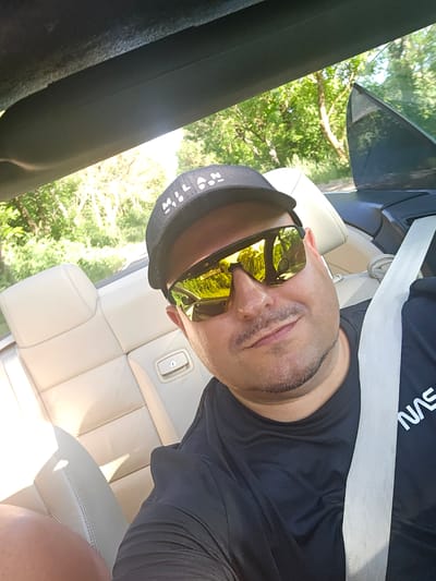 A man is sitting in a convertible, wearing a black baseball cap and sunglasses. He has a seatbelt on. The car interior is light-colored. Green trees are visible in the reflection of his sunglasses and through the car window. The location is not discernible from the image.