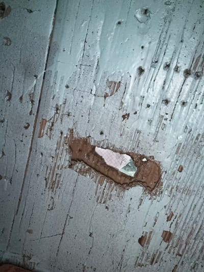 The image shows a close-up view of a weathered, pale blue wooden surface. The wood appears to be old and worn, with numerous scratches, cracks, and areas where the paint has chipped away, revealing the natural wood underneath. There are also small, dark circular indentations, possibly from nails or imperfections in the wood.
Dominating the center of the image is a roughly oval-shaped hole or damage in the wooden surface. Within this opening, a small piece of white material with a faint grid pattern is visible, possibly paper or fabric. The edges of the hole are rough and torn, exposing the raw wood and a darker, possibly dirt-filled, layer beneath.
The lighting suggests an indoor setting, possibly dimly lit, with the light source coming from above and slightly to the right, casting subtle shadows. The overall impression is one of neglect and age, with the texture of the wood and the peeling paint being the primary focus. There is no discernible text, people, or clear indication of the specific location beyond it being a wooden surface. The scene is static and conveys a sense of decay.