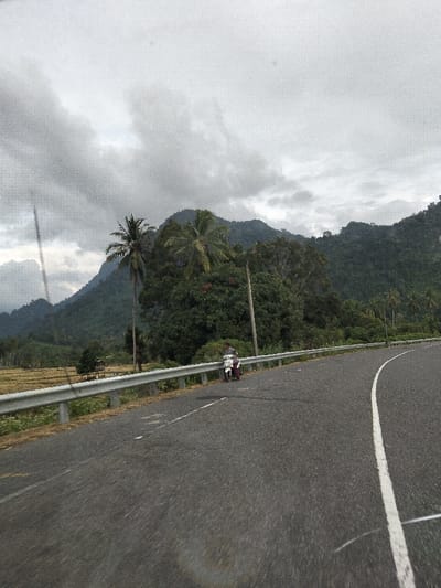 This image depicts a rural mountainous area in Keutapang, Indonesia, under an overcast sky. A paved asphalt road curves gently to the right in the foreground, featuring a solid white lane divider on its right edge. A silver metal guardrail parallels the left side of the road. On the left side of the road, adjacent to the guardrail, a male individual in a dark shirt stands next to a light-colored scooter or motorcycle, which is parked facing slightly left. Behind the guardrail and to the left of the road, a harvested or fallow field with light brown stubble is visible, containing a small, open-sided shelter with a dark roof. Several tall palm trees and dense green foliage rise beyond the field. A prominent dark utility pole stands upright behind the guardrail, positioned right of the person and scooter. The background is dominated by a lush, heavily forested mountain range. The sky is largely obscured by thick, grey clouds. The image exhibits a grainy texture and visible streaks, potentially from a camera lens or window.