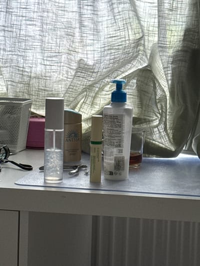 The image shows a desk with several cosmetic products and accessories, placed in front of a light-colored curtain. From left to right, there is a white mesh basket, a pink rectangular box, a clear spray bottle with a white cap, a cylindrical bottle with a tan label that reads "ANESSA", a small tube with green and white labeling, and a clear glass with a dark liquid. A pair of black-handled tweezers and a silver nail clipper are also visible on the surface. The lighting suggests it is daytime, with sunlight filtering through the curtain. The desk appears to be a vanity or dressing table. The location of Коробицыно, Russia, is not discernible from the image content.