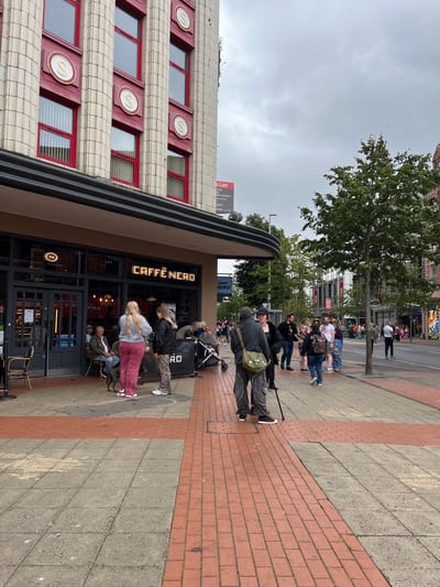 The image shows a view of a street scene in Belfast City District, United Kingdom, under an overcast sky. In the foreground, a Caffe Nero coffee shop is visible, with several people standing and sitting outside. Two women are standing in front of the Caffe Nero entrance, one wearing pink trousers and a grey top, the other in a black jacket and leopard print trousers. A man is seated outside the cafe, and a dog is standing next to him. Further down the sidewalk, a line of people is visible, and a tree grows by the roadside. The building behind the cafe has red-framed windows with circular gold emblems. The street is paved with grey and red bricks, and there is a road with traffic in the background. The overall atmosphere appears to be casual and public, with people engaged in everyday activities.
